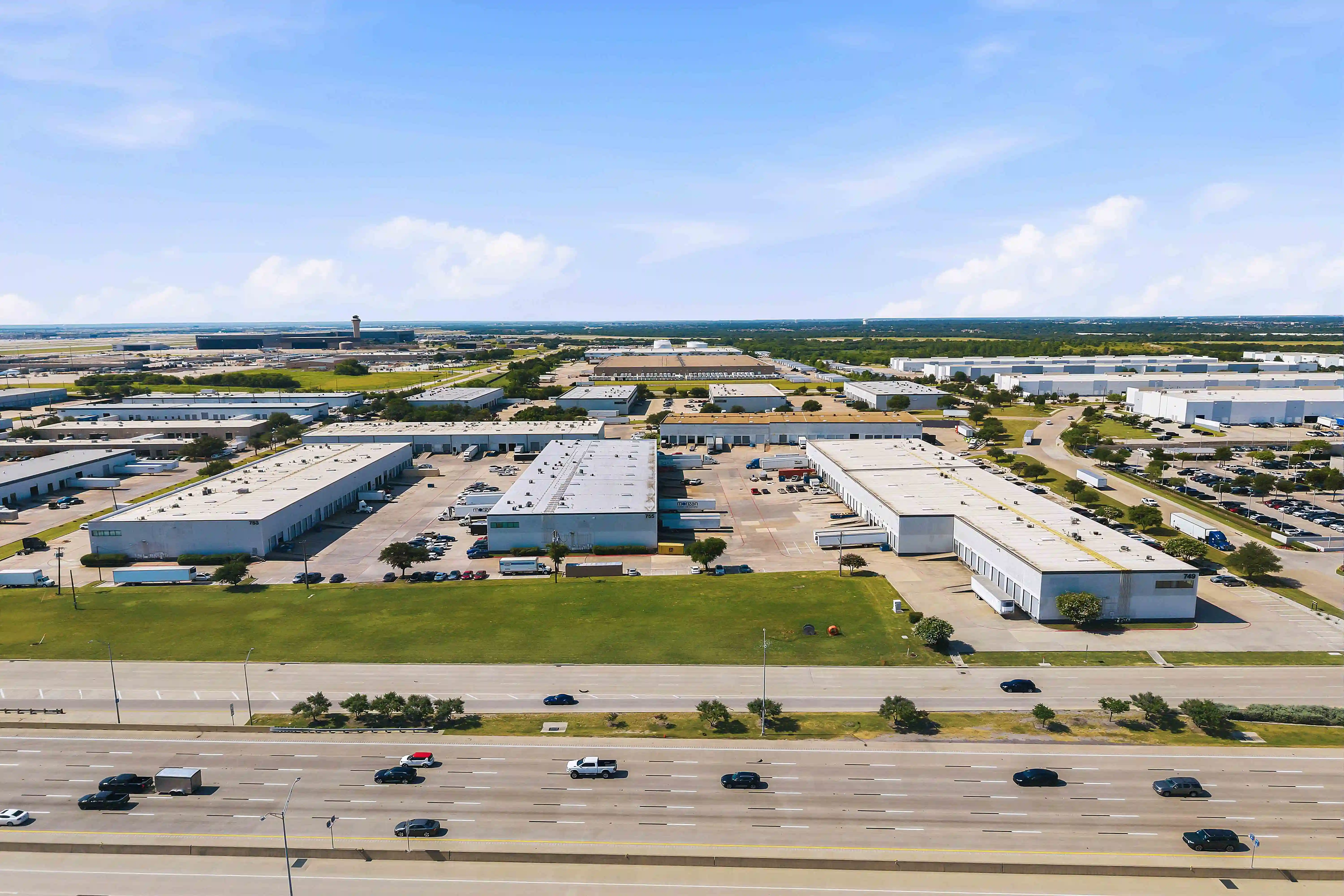 A high aerial view captures warehouse clusters, truck courts, green setbacks, and a multilane freeway crossing the foreground.